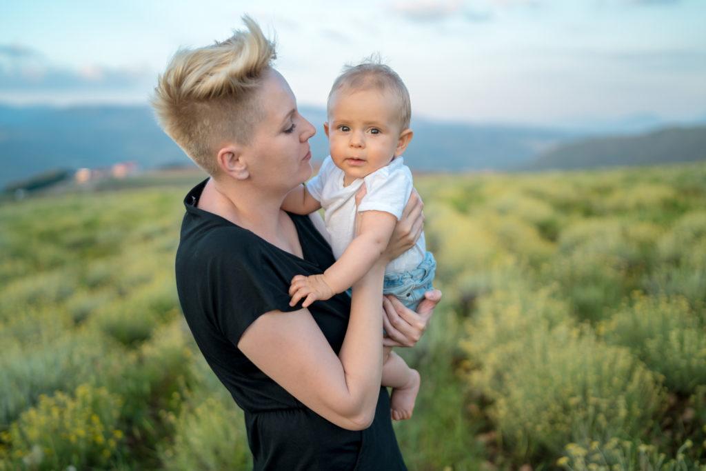 Portrait of young mother holding little baby in hands standing in bright yellow field of flowers