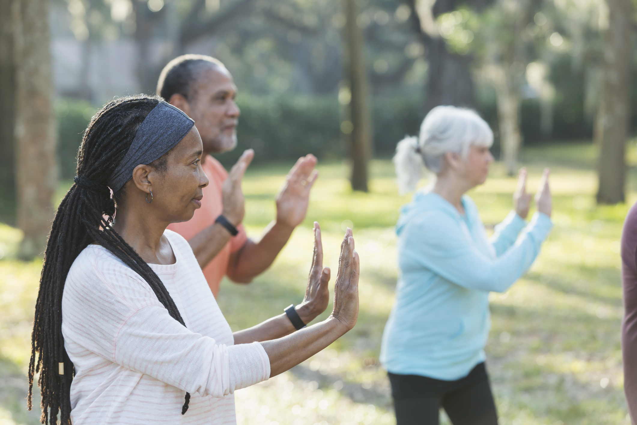 A group of three multi-ethnic seniors taking an exercise class in the park. They are practicing tai chi, standing with their hands raised. The focus is on the Africam-American woman with braided hair standing in the foreground.