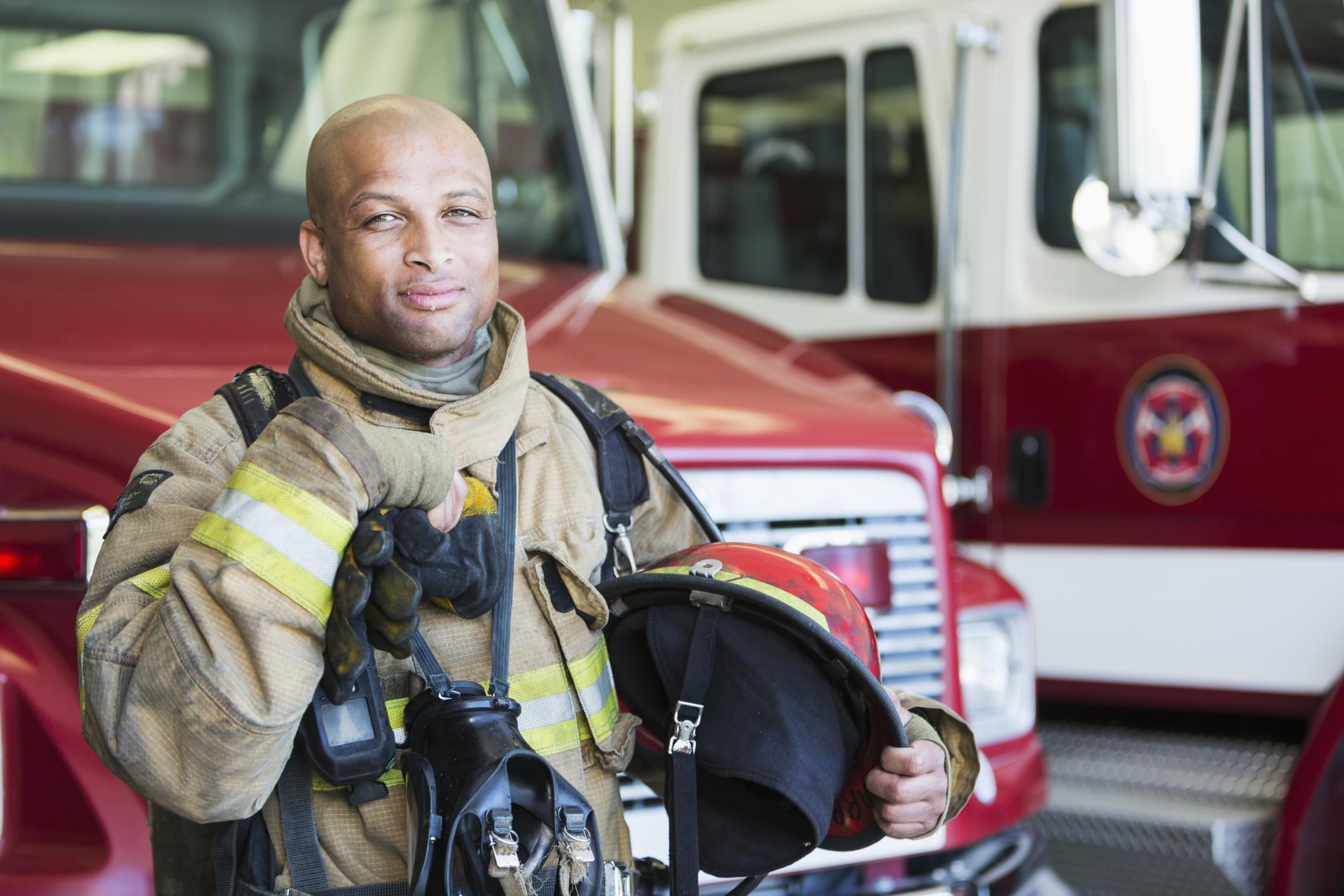 Portrait of an African American fireman standing in front of a fire engine parked at the station. He is serious and confident, wearing protective suit, holding gloves and a helmet. He is ready to respond to an emergency.