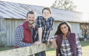 A family with a teenage son standing outside a farm building, leaning on a wooden fence, smiling at the camera. It is bright and sunny.