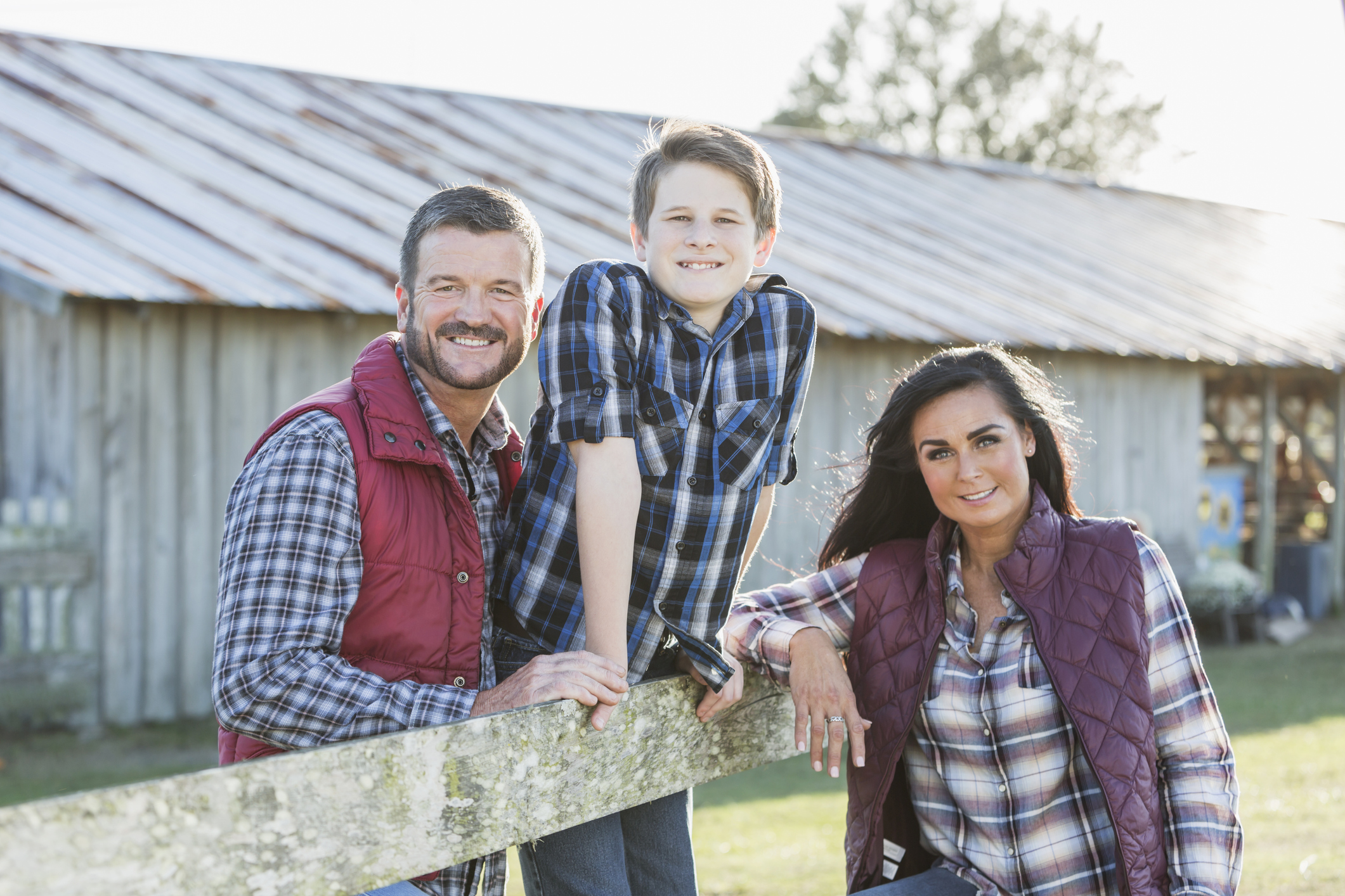A family with a teenage son standing outside a farm building, leaning on a wooden fence, smiling at the camera. It is bright and sunny.