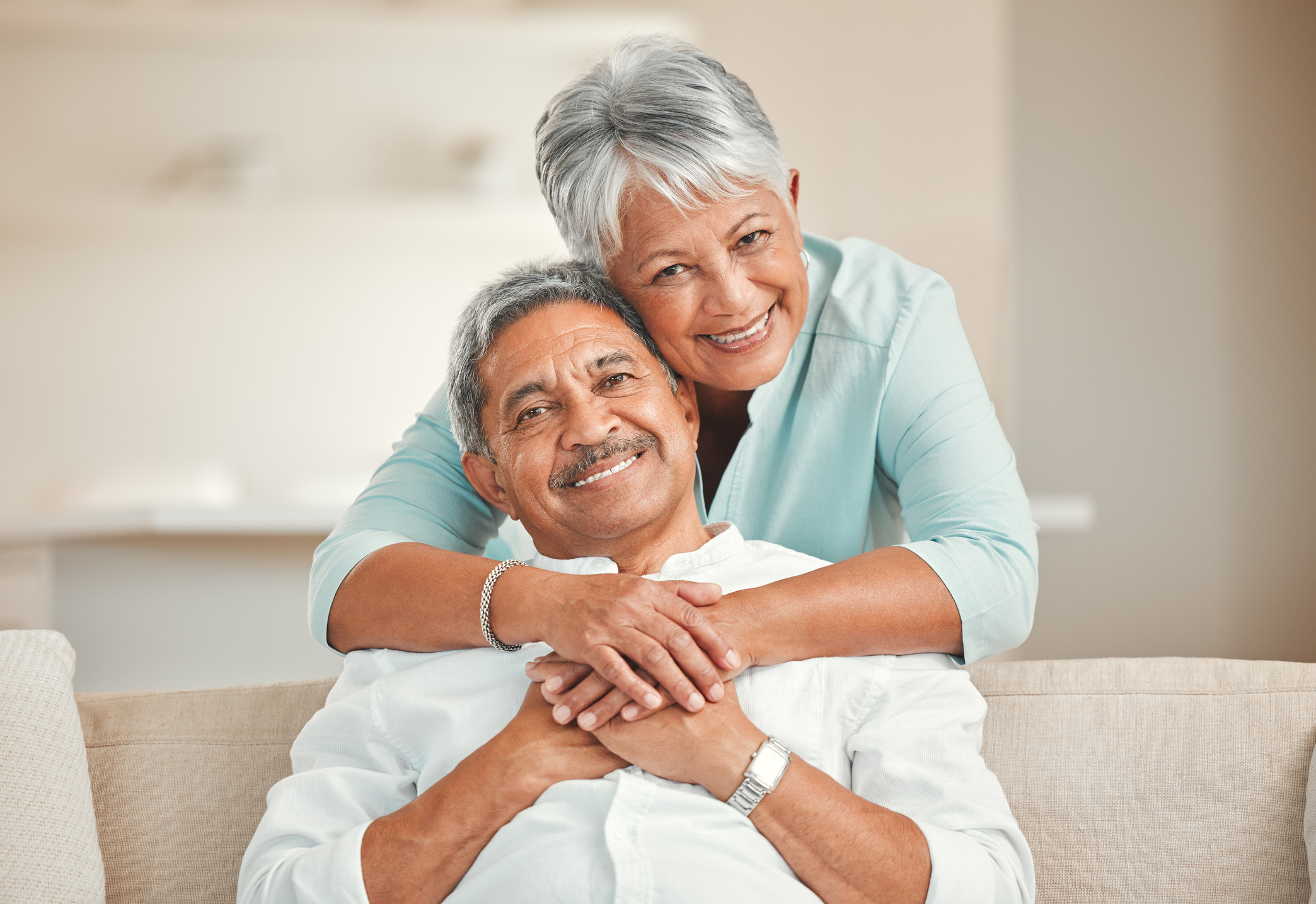 Older woman smiling and putting her arms around the shoulders of an older man sitting on the couch and smiling