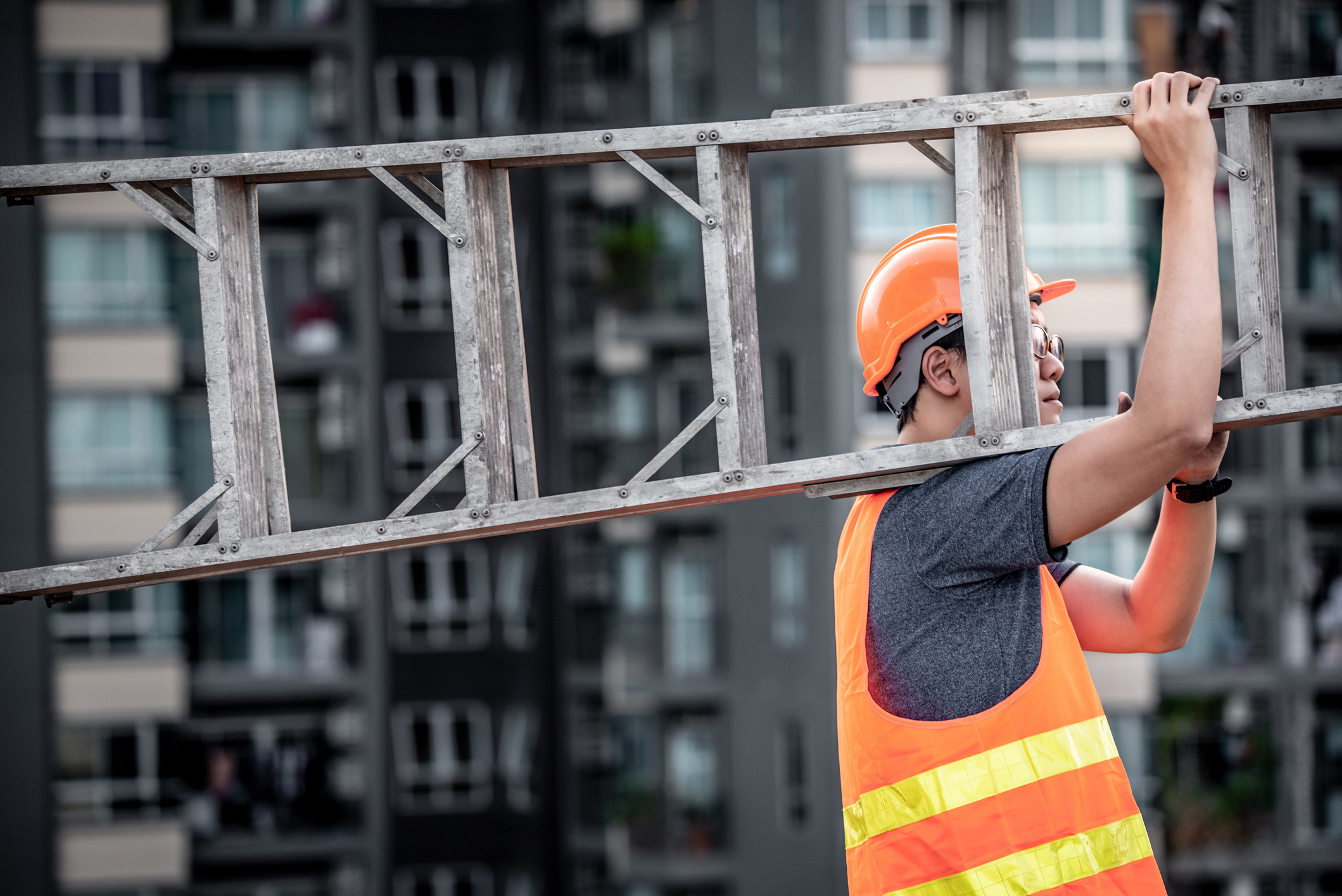 Maintenance worker with orange safety helmet and vest carries a ladder.