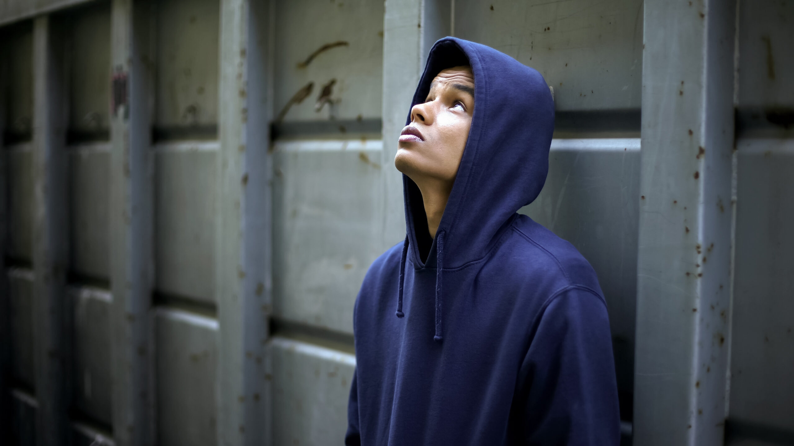 Sad, teen boy in a blue hoodie stands by a wall of a building and looks up