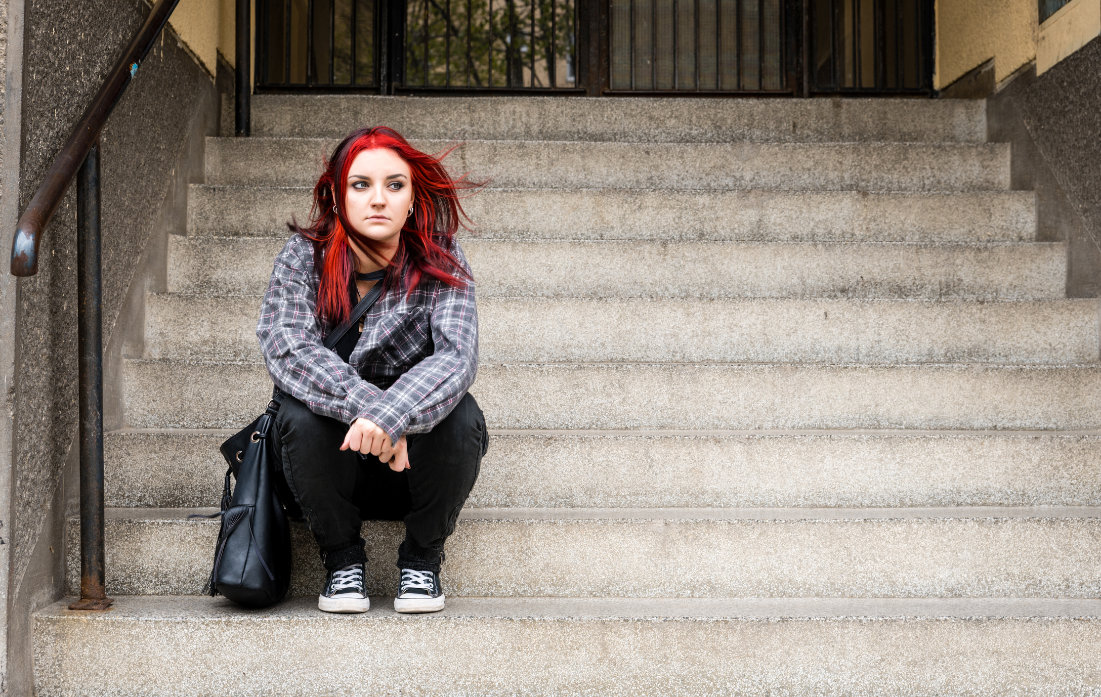 A teen girl sits on outdoor stairs with a bag.