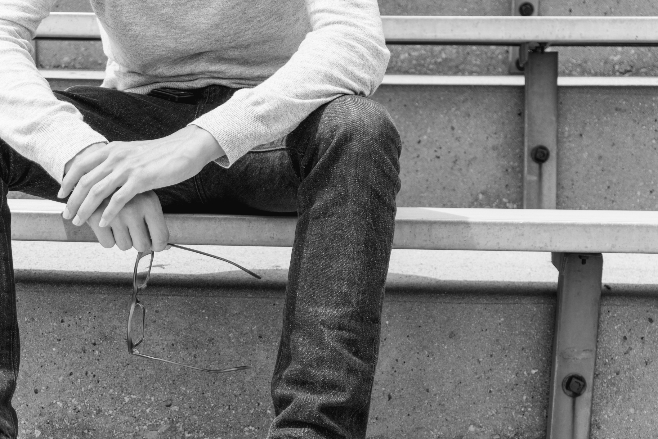 Teen boy sitting on an outdoor bench holding a pair of eye glasses. Photo does not show face.