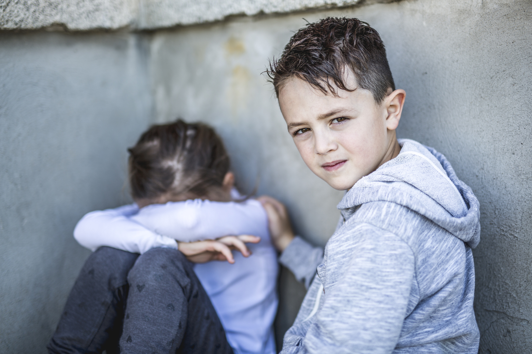 Two young sad-looking children sitting by an outdoor gray wall.. Boy is comforting the girl while looking at the camera. Girl is sitting with her head in her arms.