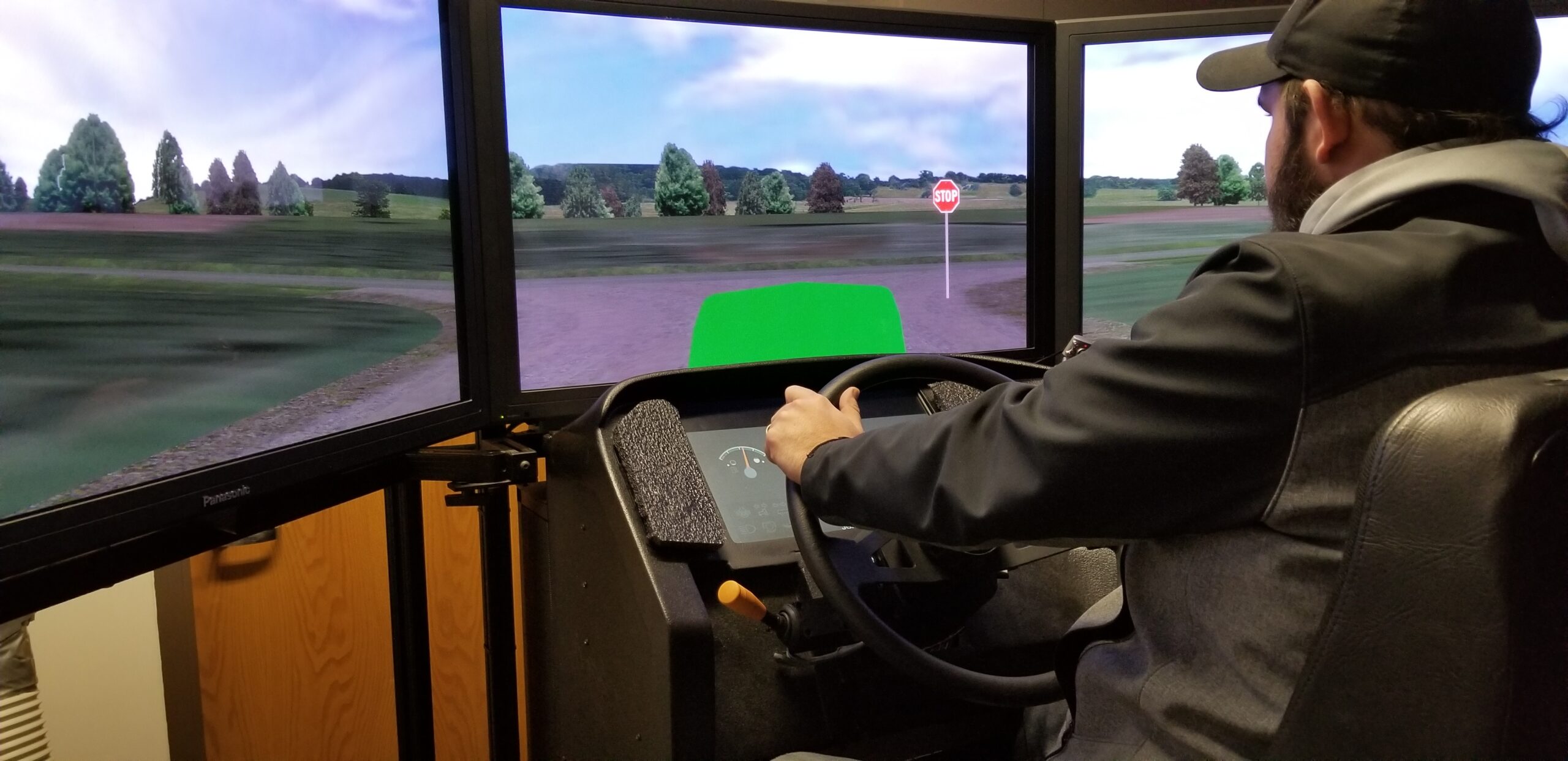 a man drives a virtual reality tractor simulator. he is at the wheel and a virtual rural road is the view out the window.