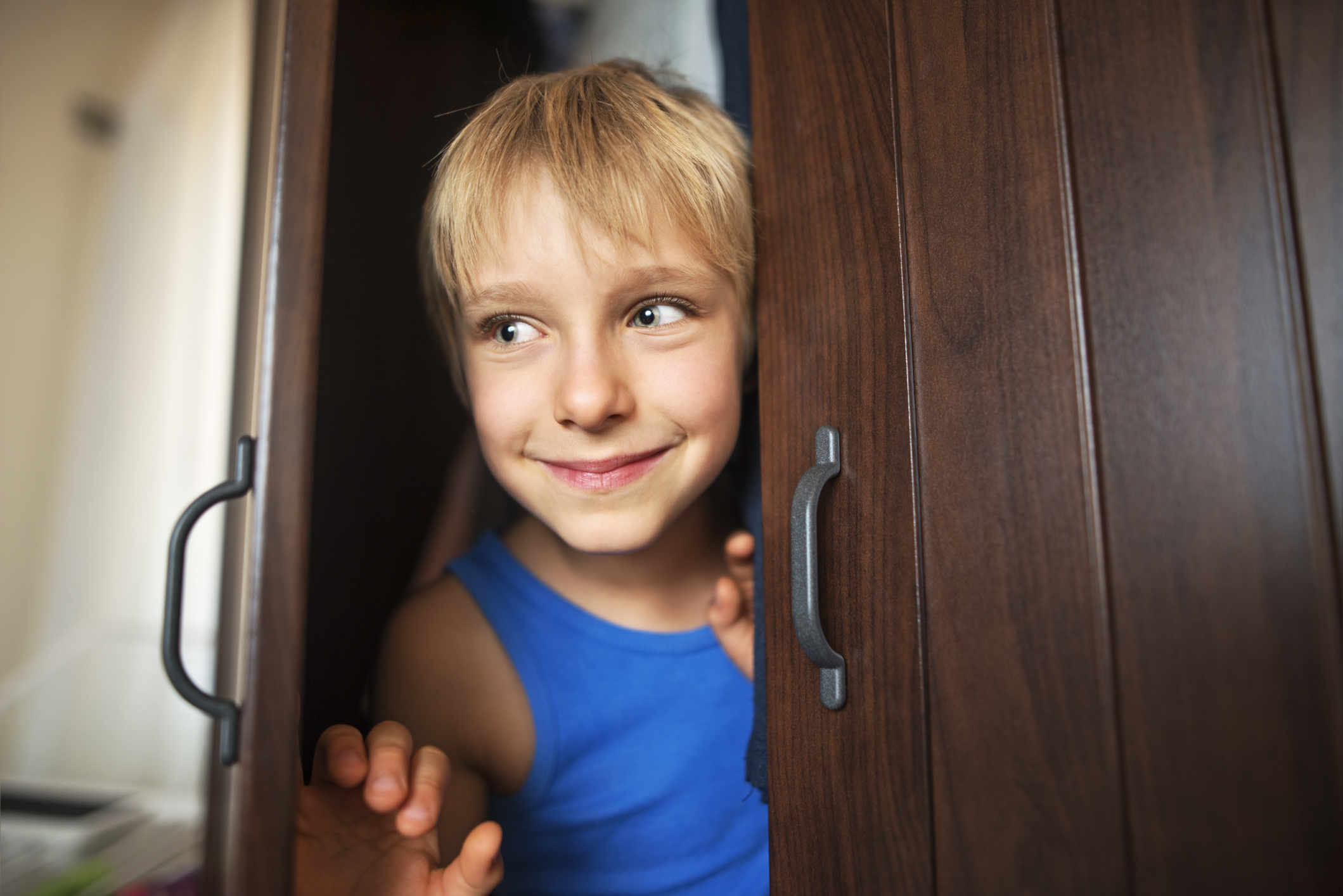 Little boy is playing hide and seek. The boy aged 7 is hiding in a closet. He is opening the door and smiling.