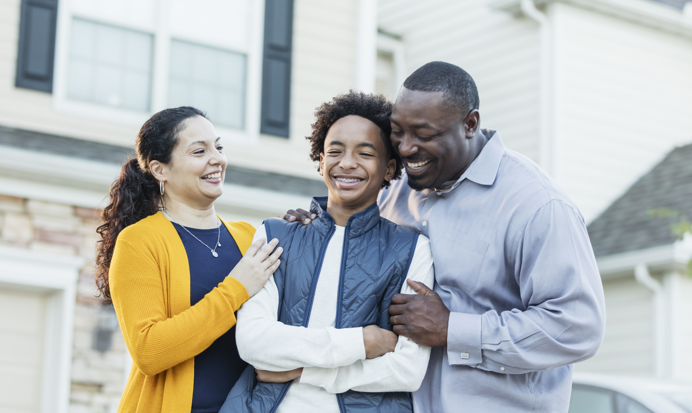 A family of three smiling in front of a house: A teen boy in the middle of his parents 