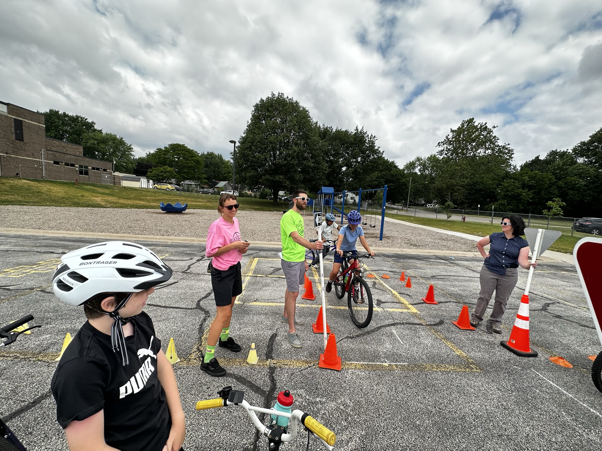 Bicycle education program in a parking lot at a school. A few children on bikes biking around cones and research staff helping