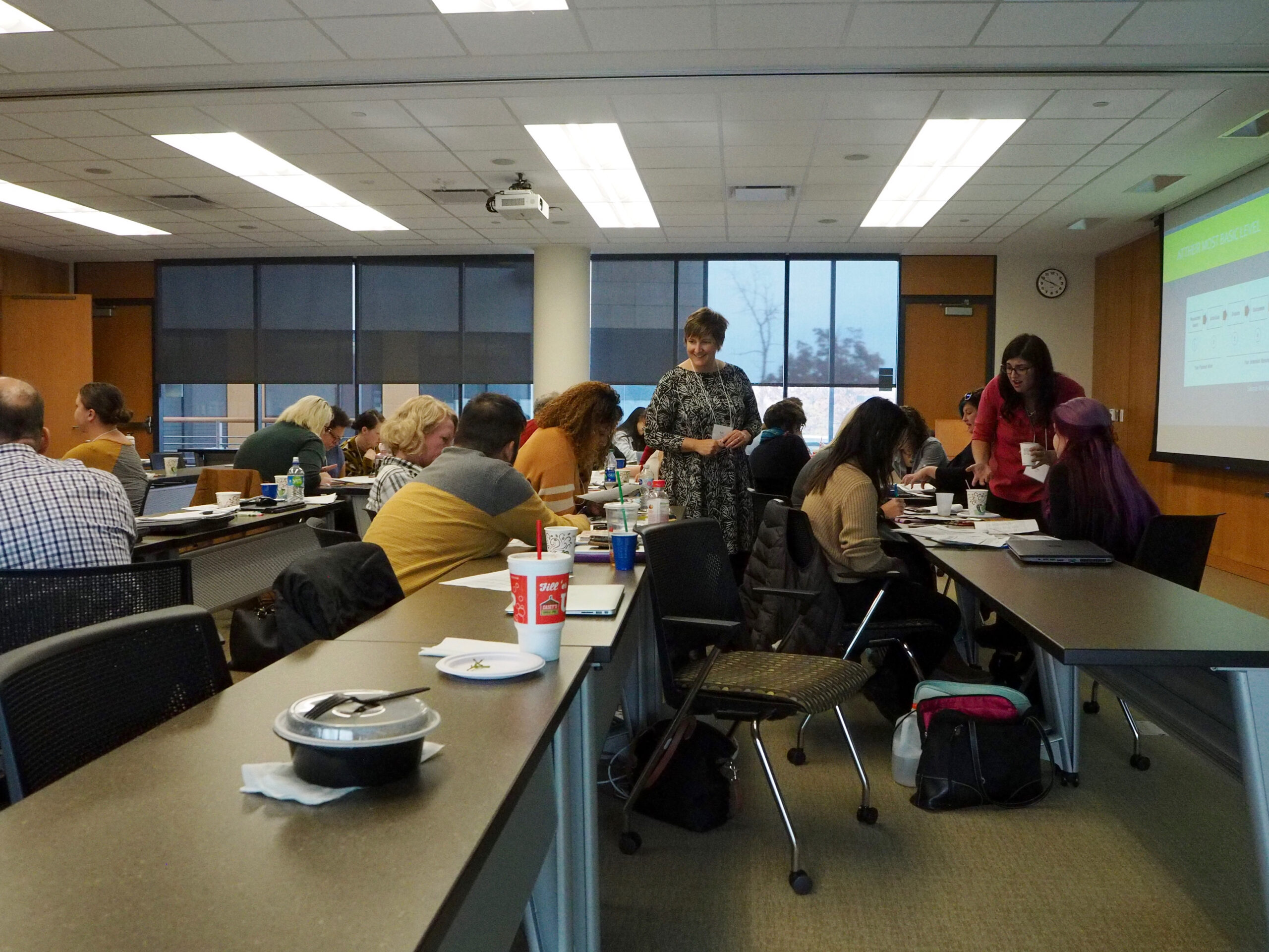 About a dozen people sit at long desks with 2 instructors standing