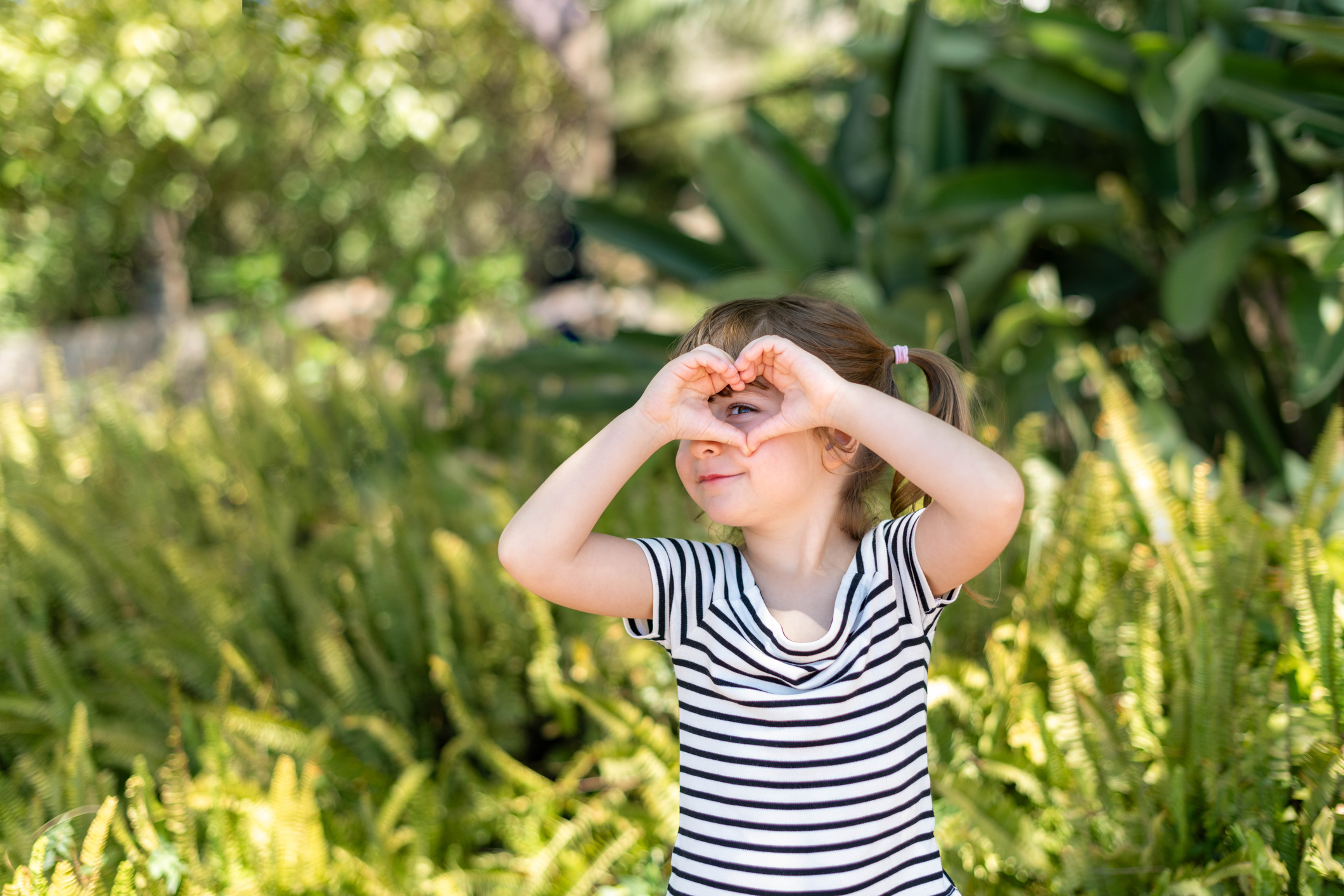 Portrait of girl with her hand in the shape of a heart on her eye, on green nature background