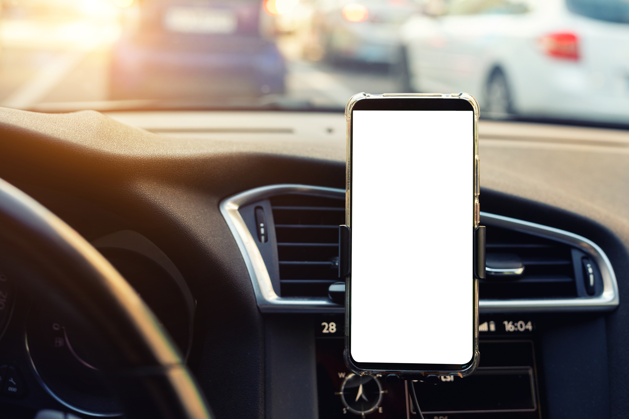 Interior of a car with a smartphone mounted on the dashboard displaying a blank white screen. The car's air vents and part of the steering wheel are visible. Outside the windshield, traffic is seen with several cars on the road under bright sunlight.