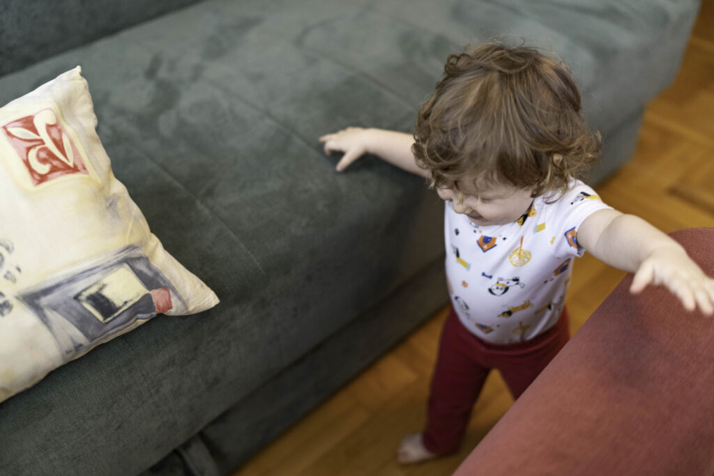 Baby walking between a couch and another piece of furniture and holding onto the sides and smiling