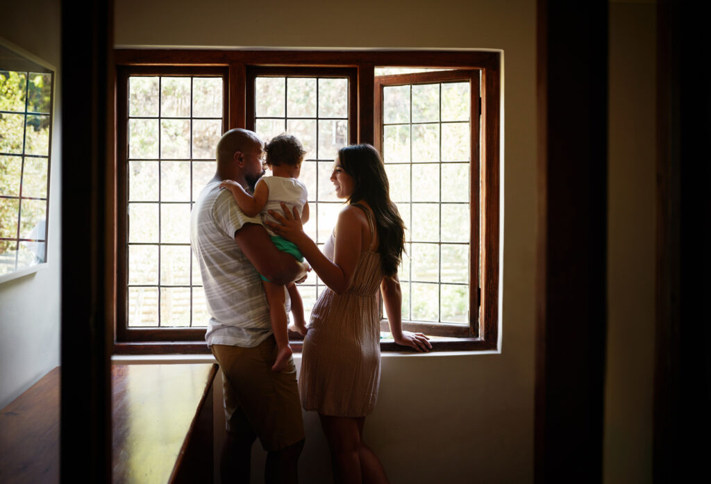 A man holds a young child next to a smiling woman inside a home. They are looking out a large window.