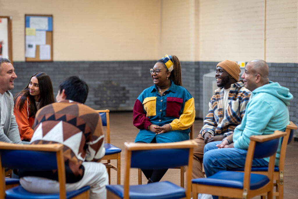 Six people sit in chairs in a circle and smile and laugh. They are in a community center and dressed in casual clothing.