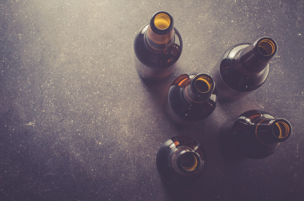 Five empty brown glass bottles arranged on a dark, textured surface. The bottles are positioned in a scattered manner, with their openings facing upwards. 
