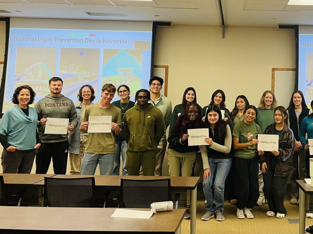 A group of over 15 people smiling in a classroom in front of a screen. A few are holding signs that say #BeInjuryFree.