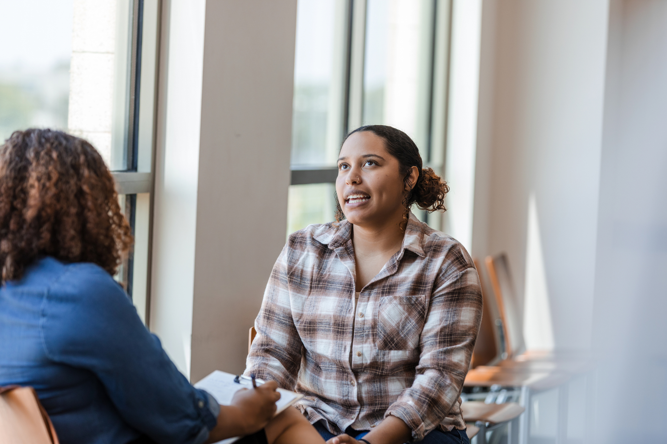 Young woman talks to another woman writing on a clipboard. Both are sitting down.
