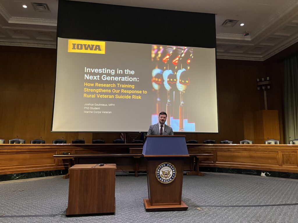 A man stands at a podium with a screen behind him that says: Investing in the Next Generation: How Research Training Strengthens Our response to Rural Veteran Suicide Risk